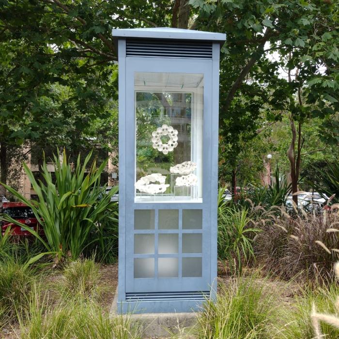 Display case with abstract white sculptures resembling interconnected rings and shapes, set against a backdrop of greenery.