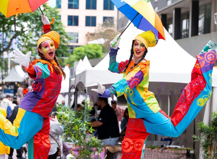 Two stilt walkers dressed in vibrant, multicoloured costumes perform at an outdoor event in an urban setting. They are wearing bright face paint, exaggerated expressions, and matching yellow headpieces. Each holds a rainbow-coloured umbrella, adding to th