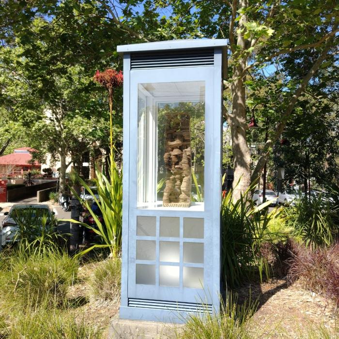 A tall, intricately carved wooden sculpture displayed inside a glass enclosure, placed in an outdoor setting with trees and plants visible in the background.