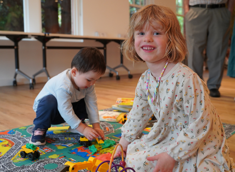 Two young children playing with toy cars and trucks on a playmat. One of them is looking directly at the camera and smiling.