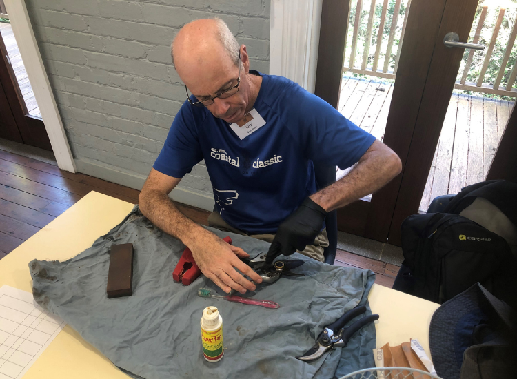 A man sitting at a table, wearing a glove and with various tools, fixing a pair of secateurs.