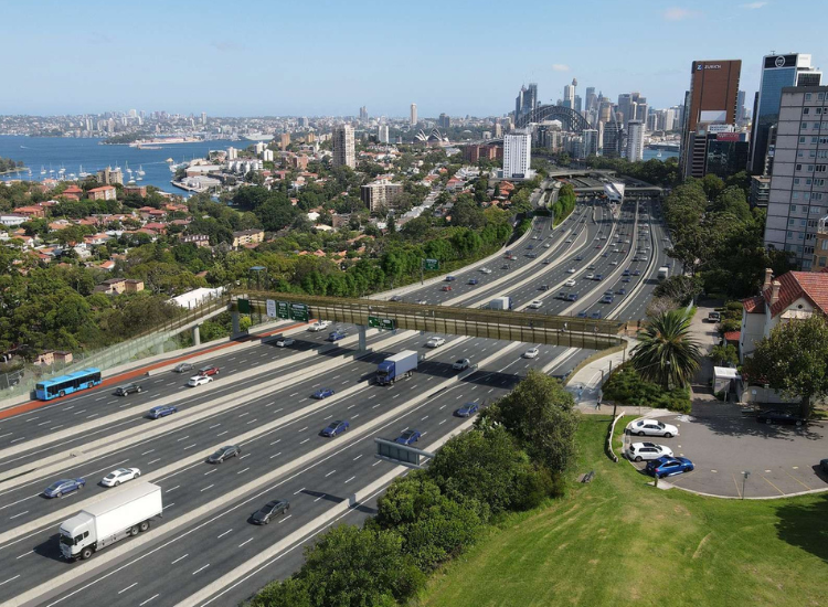 An impression of what the upgraded Ridge Street shared access bridge will look like once completed: aerial shot showing the pedestrian bridge across a freeway, with Sydney Harbour and the Sydney and North Sydney CBDs in the background.