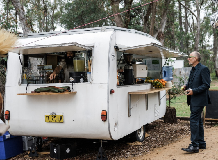 A white caravan with a cafe set up inside, with bushland in the background and a customer standing outside.