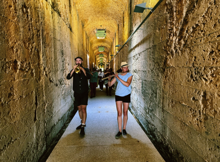 A man playing a trumpet and a woman playing a flute, walking through a brightly lit tunnel, with a group of people behind them.