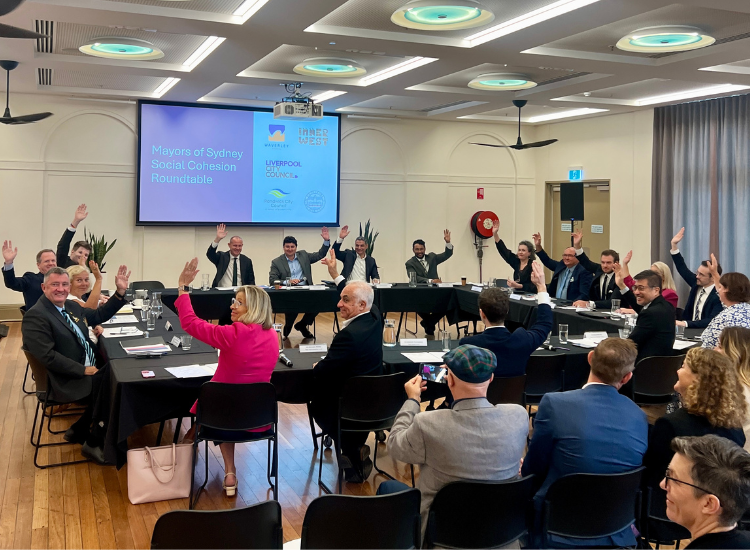 A group of around 20 mayors sitting at tables arranged in a square shape, holding their hands in the air to vote 'yes'.