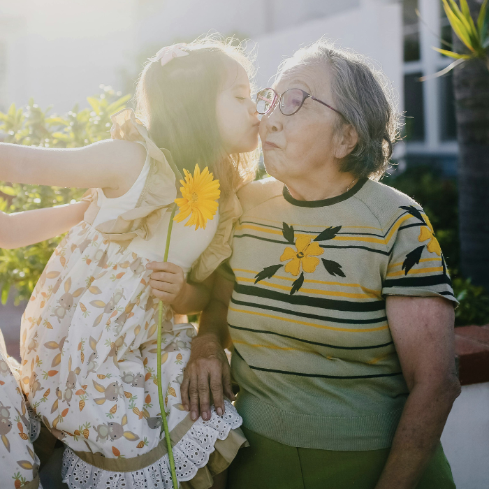 A young girl, holding a flower, and kissing an older woman on the cheek.