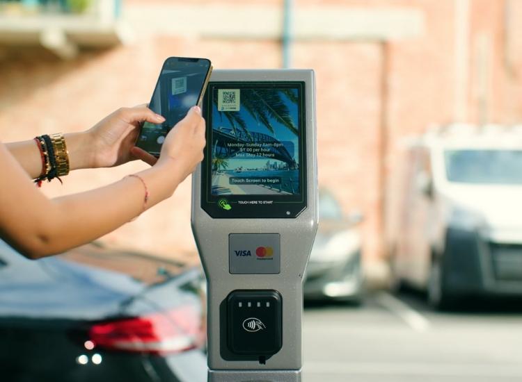 modern touch screen parking meter with cars behind it and person holding their phone infront