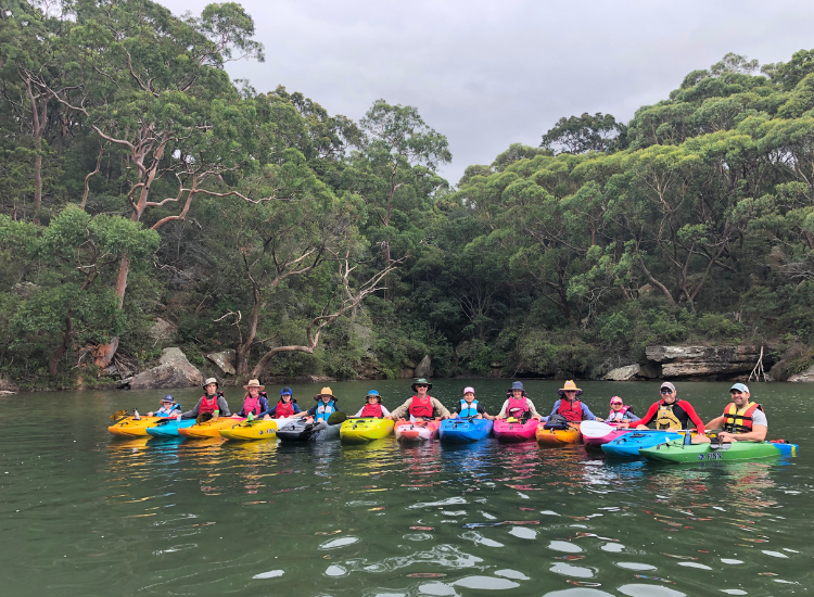 A group of people in colourful kayaks on the water in front of native bushland.