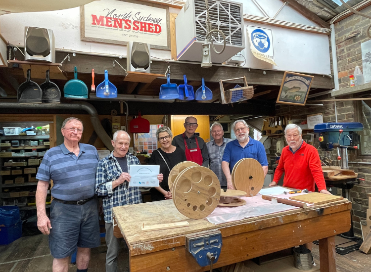 A group of six men and one woman, standing behind a table in a shed workspace. A sign above them reads &#039;North Sydney Men&#039;s Shed&#039;. On the table are three circular wooden devices with holes in them. One man holds a certificate.