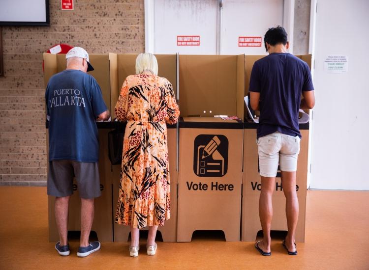 three people with their back to camera standing at a cardboard polling booth