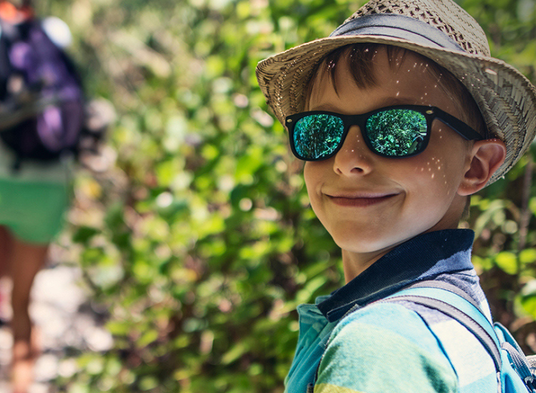 A young boy wearing a hat, sunglasses and t-shirt, smiling at the camera with green bushland in the background